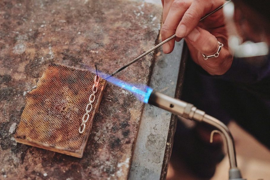 Eine Hand hält einen Brenner, der eine Kette auf einer Metallplatte erhitzt.
