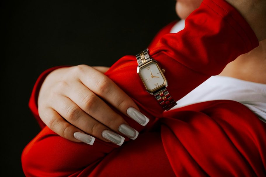Eine Hand mit langen Nägeln trägt eine elegante Uhr und berührt einen roten Stoff.