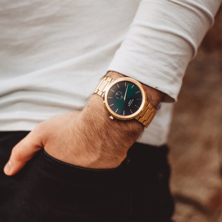 Hand mit Armbanduhr in Gold und grünem Ziffernblatt, Baumwollshirt und schwarze Hose.