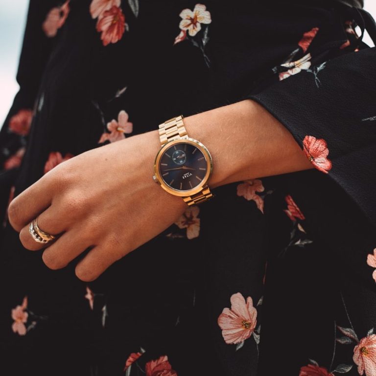 Armband mit goldenem Gehäuse und schwarzem Ziffernblatt an einer blumigen Kleidung.
