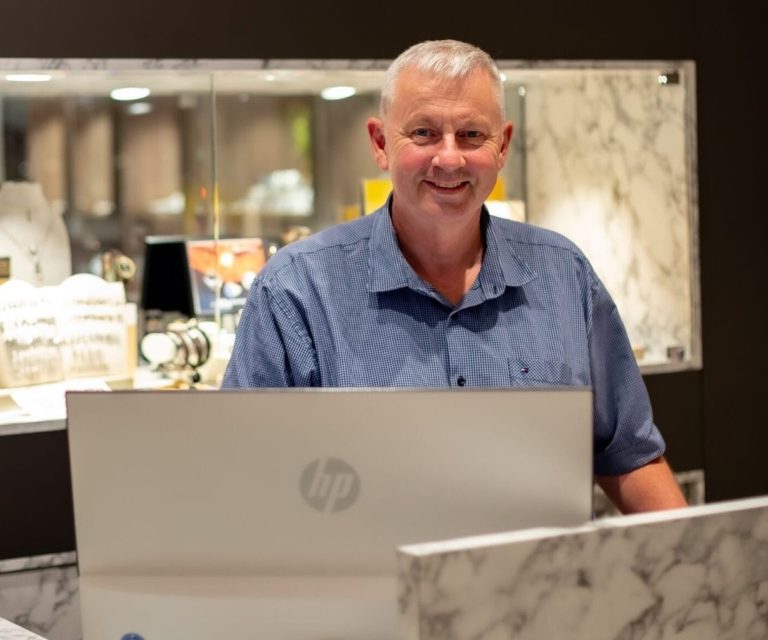 Mann am Schalter mit Laptop, lächelt freundlich. Schmuckauslage im Hintergrund.