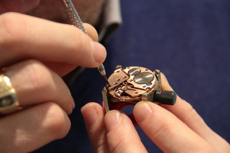 Eine Hand hält eine detailreiche Uhr, während die andere sie mit einem Werkzeug bearbeitet.